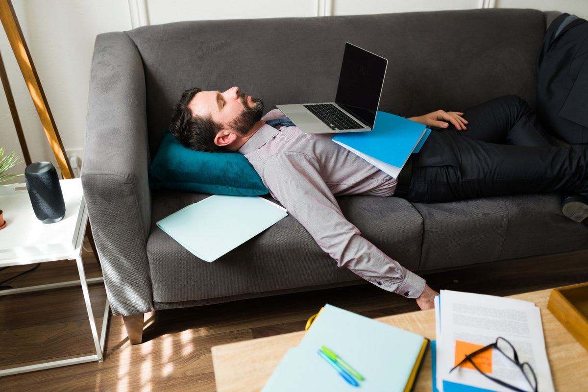 A tired man asleep on a sofa with a laptop and folders on his chest