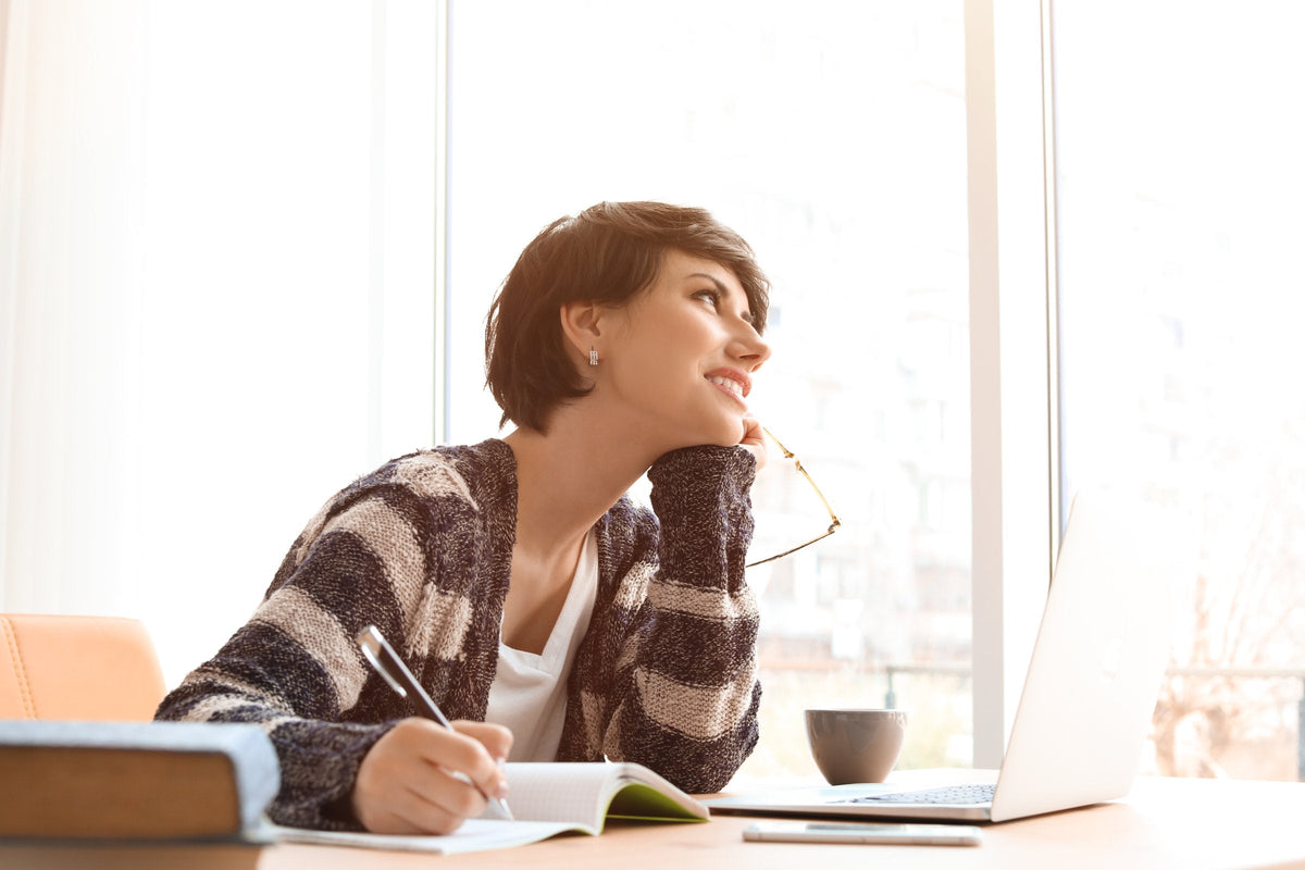 A woman at a desk by the window, pen on her notebook, drifting off in thought